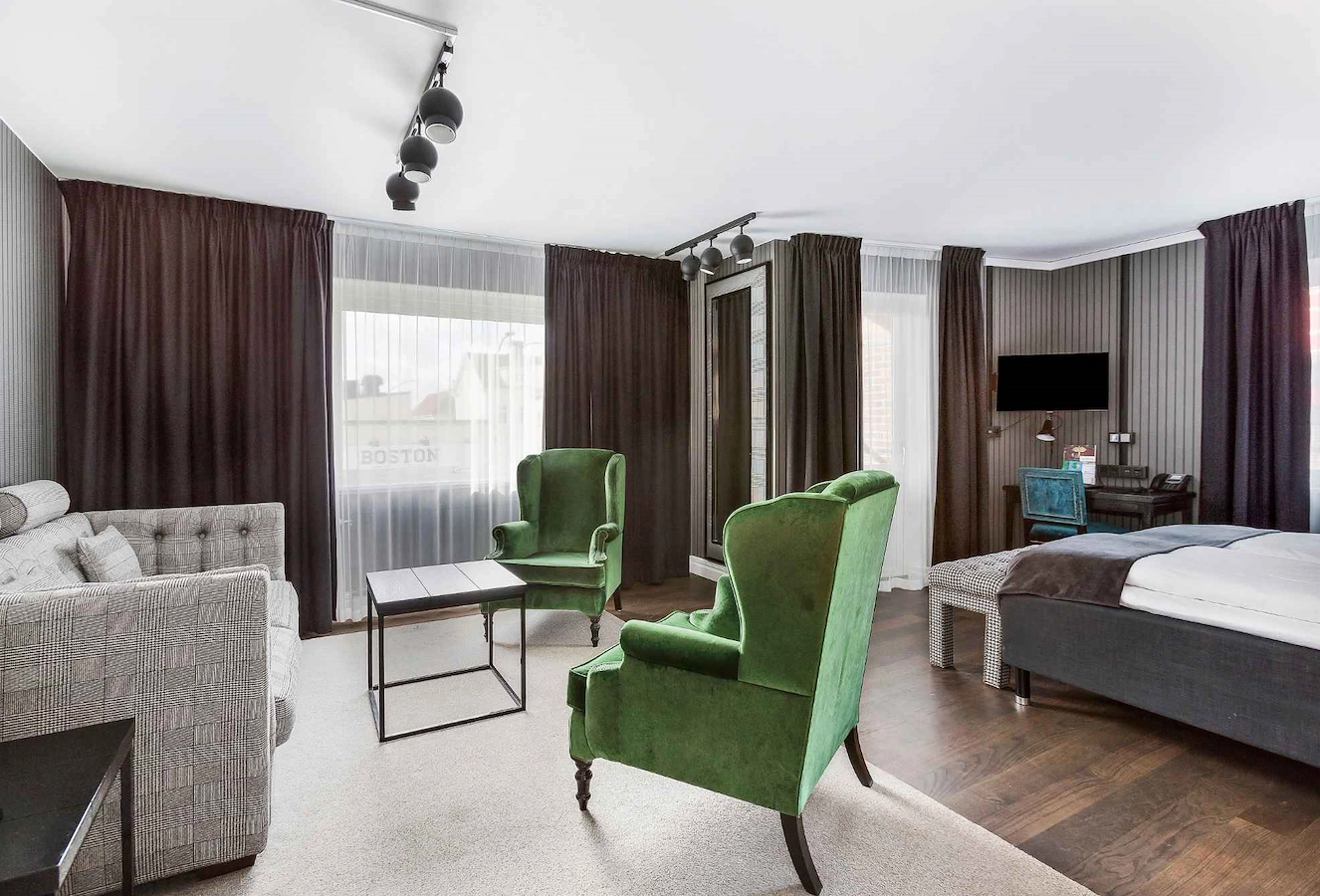 Rymligt familjerum superior på Vimmerby Stadshotell med dubbelsäng, bäddsoffa och en loungedel med två gröna sammetsfåtöljer och bord. Flera fönster och balkongdörr släpper in ljus i det mörkt inredda rummet med unika detaljer.