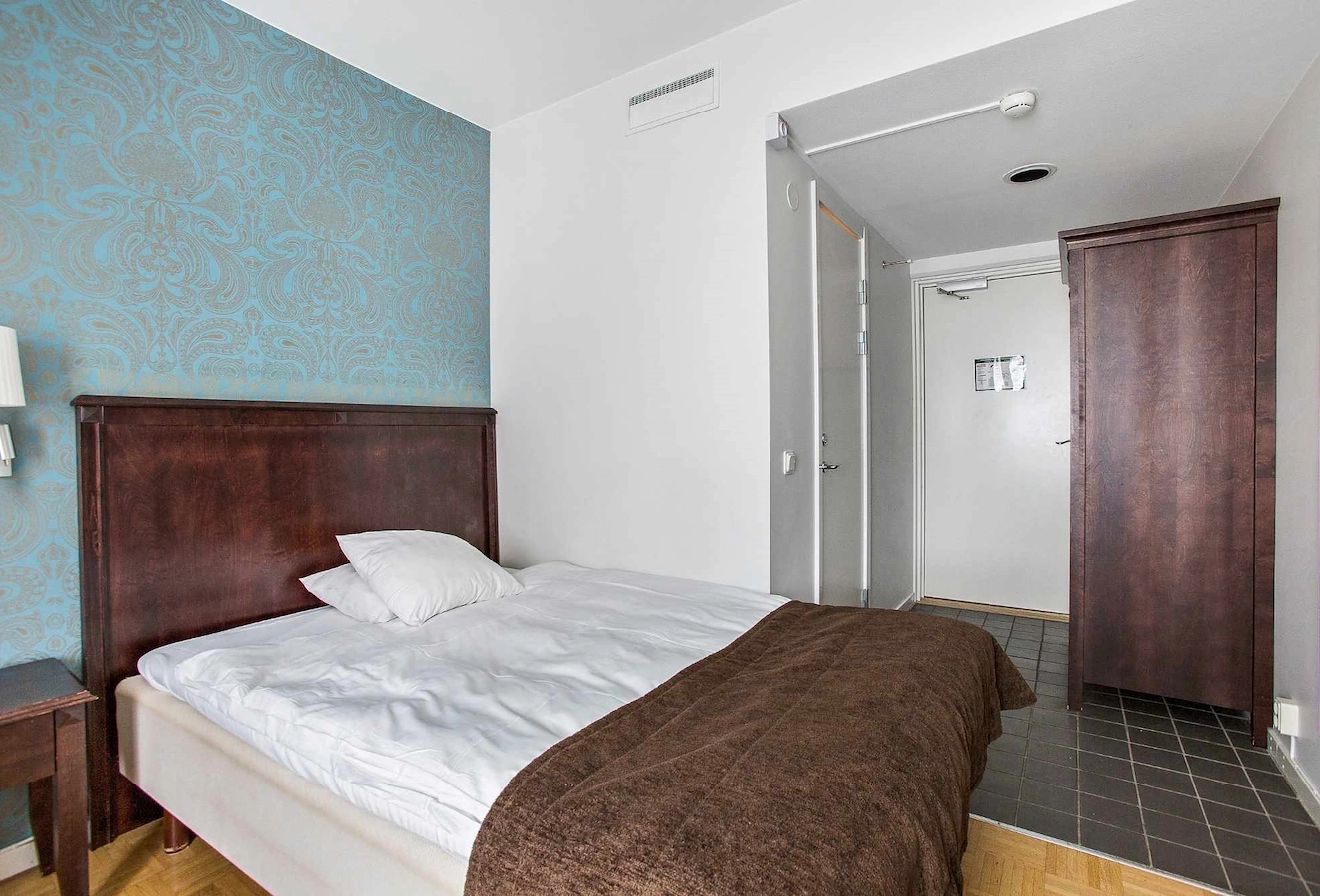 Economy dubbelrum på Vimmerby Stadshotell – 140 cm säng, turkos fondvägg och mörka trädetaljer i ett klassiskt hotellrum. Klinker vid entrén, rent, fräscht och enkelt boende för två personer eller en person som vill ha en större säng. 