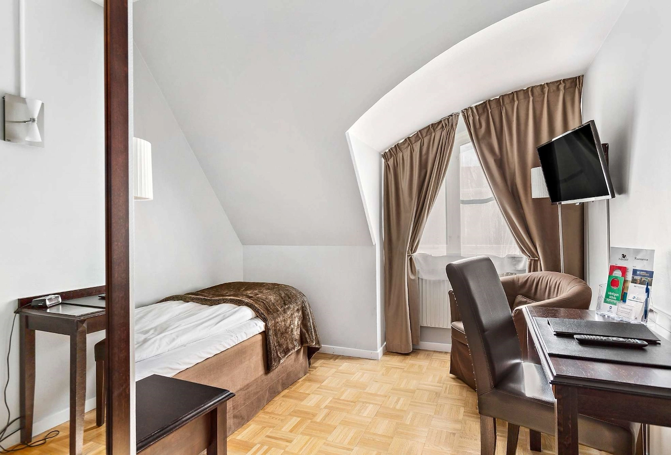 Stilrent och lugnt enkelrum på Vimmerby Stadshotell – med 90 cm enkelsäng, skrivbord, fåtölj och nattduksbord. Ljusa väggar, mörka trädetaljer och ett fönster med skira gardiner och mörkläggning. Perfekt för egen tid eller affärsresan.