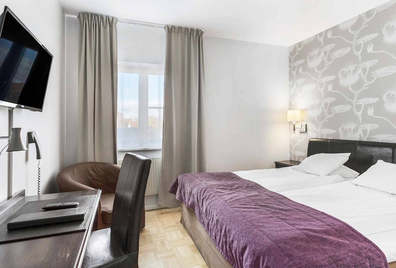Klassiskt dubbelrum på Vimmerby Stadshotell – inrett i ljusa färger med beige fondvägg, krispigt vita lakan, skrivbord, fåtölj och TV. Mörka trädetaljer ger rummet en varm och tidlös känsla.