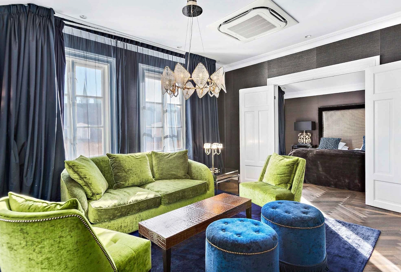 Svit Direktören på Vimmerby Stadshotell – lyxigt allrum med grön sammetsoffa, matchande fåtöljer, blå puffar och rikligt med textilier. Modern kristallkrona i taket och sovrum med dubbelsäng syns bakom eleganta dubbeldörrar.